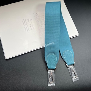 May include: A teal blue woven fabric bag strap featuring textured leather end tabs with white contrast stitching and silver-toned metal swivel lobster claw clasps at both ends. The strap is displayed alongside a white card with printed text.