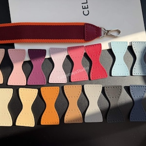 May include: A collection of small, hourglass-shaped leather patches in various colors including pink, red, blue, orange, and grey, each featuring pre-punched stitching holes. A long, two-toned maroon and orange fabric strap with a metal clasp is included.