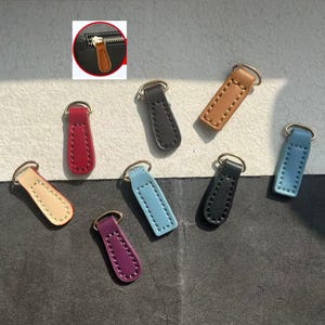 May include: A collection of leather zipper pulls in various colors, including red, black, tan, and blue. Each pull has a metal ring for attachment. The pulls are designed for use on bags, jackets, or other items with zippers.