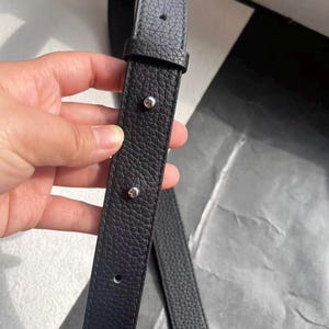 Op de afbeelding: Close-up van een zwarte leren riem met een getextureerd oppervlak. De riem heeft zilverkleurige klinknagels en stiksels. De riem is waarschijnlijk voor een tas of portemonnee.