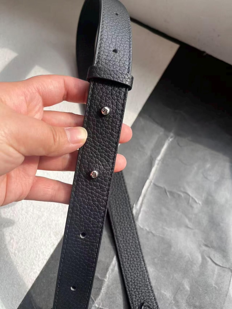Op de afbeelding: Close-up van een zwarte leren riem met een getextureerd oppervlak. De riem heeft zilverkleurige klinknagels en stiksels. De riem is waarschijnlijk voor een tas of portemonnee.