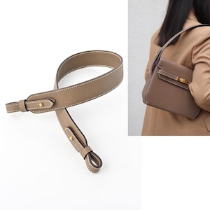 Puede incluir: Una correa de bolso de mano de cuero color topo con costuras blancas y herrajes dorados. La correa se muestra junto a un bolso a juego. La correa está diseñada para sujetarse a un bolso.