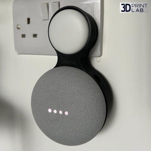 Puede incluir: Un soporte de pared negro impreso en 3D que sostiene un altavoz inteligente Google Nest Mini. El altavoz es gris con cuatro puntos iluminados, y el soporte está fijado a una pared blanca cerca de una toma de corriente. El logotipo "3D PRINT LAB" es visible.