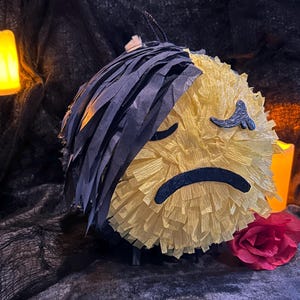 May include: A round piñata with a sad face design, made of yellow crepe paper and black fringe. A red rose is placed near the piñata. Two lit candles are in the background, creating a moody atmosphere.