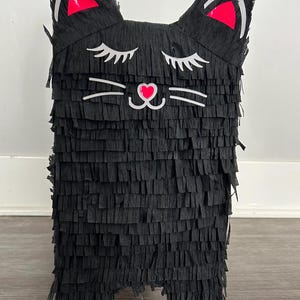 May include: Black cat piñata with a fringed body and cat face details. The cat's ears have red accents, and the face features white whiskers and closed eyes. A heart shape is in the center of the face.