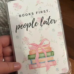 May include: A clear phone case with a floral design and the text "BOOKS FIRST, people later." The case displays a stack of books tied with a pink ribbon. The books are in shades of orange, green, and blue.