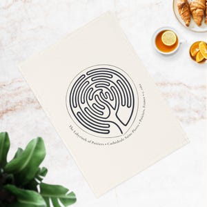 May include: A rectangular off-white tea towel featuring a centred black circular labyrinth design. The text surrounding the labyrinth reads: The Labyrinth of Poitiers • Cathédrale Saint-Pierre • Poitiers, France • c. 1162. The fabric is plain.