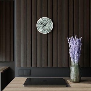 Puede incluir: Un reloj de pared circular con un patrón de laberinto verde oscuro sobre fondo blanco y manecillas negras. El reloj está montado en una pared de listones verticales de color marrón oscuro sobre un portátil negro y un jarrón con lavanda.