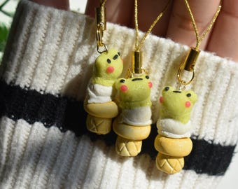 May include: Three handmade frog-shaped charms. Each charm features a light green frog with pink cheeks, a white layer, and a yellow ice cream cone base. The charms are attached to gold-colored cords.