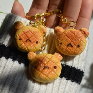 May include: Three handmade bear-shaped keychains. Each keychain features a yellow-orange bear with a waffle pattern and a small, sad face. The keychains are attached to gold-colored chains and rings. The background includes a white and black striped sweater.