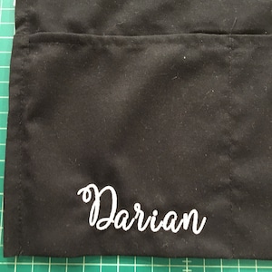 May include: Black fabric apron with a white embroidered name "Darian" on the bottom.