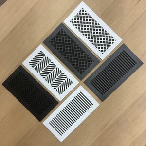 Op de afbeelding: Een assortiment rechthoekige ventilatieroosters in verschillende ontwerpen en kleuren, waaronder wit, zwart en grijs. De patronen omvatten ruitvormige rasters, parallelle lijnen en chevronvormen. Deze decoratieve roosters zijn gerangschikt op een licht houten oppervlak.