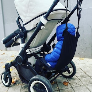 Può includere: Un'amaca nera per bambini sospesa a un passeggino bianco e nero. Un bambino indossa una giacca blu imbottita. Il passeggino ha ruote grandi. Lo sfondo è un muro grigio e una superficie pavimentata.