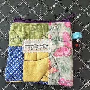 May include: A small quilted zippered pouch with a patchwork design. The pouch is made of green, yellow, blue, and pink fabric with a floral print. The pouch has a label that says "Quarantine Quilter Lockdown Loot Bag".
