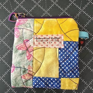 May include: A small quilted zippered pouch with a patchwork design featuring butterflies, yellow, blue, and white fabrics. The pouch has a label that reads "Quarantine Quilter Lockdown Loot Bag".