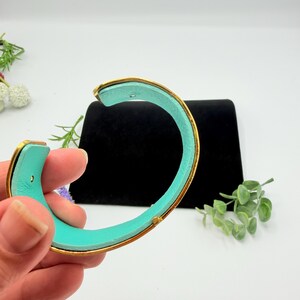 May include: A C-shaped cuff bracelet featuring a bright turquoise interior lining and a metallic gold-toned exterior frame. The bracelet has a smooth, curved design with a visible opening, showcasing a vibrant contrast between the inner colour and the outer gold-toned metal finish.