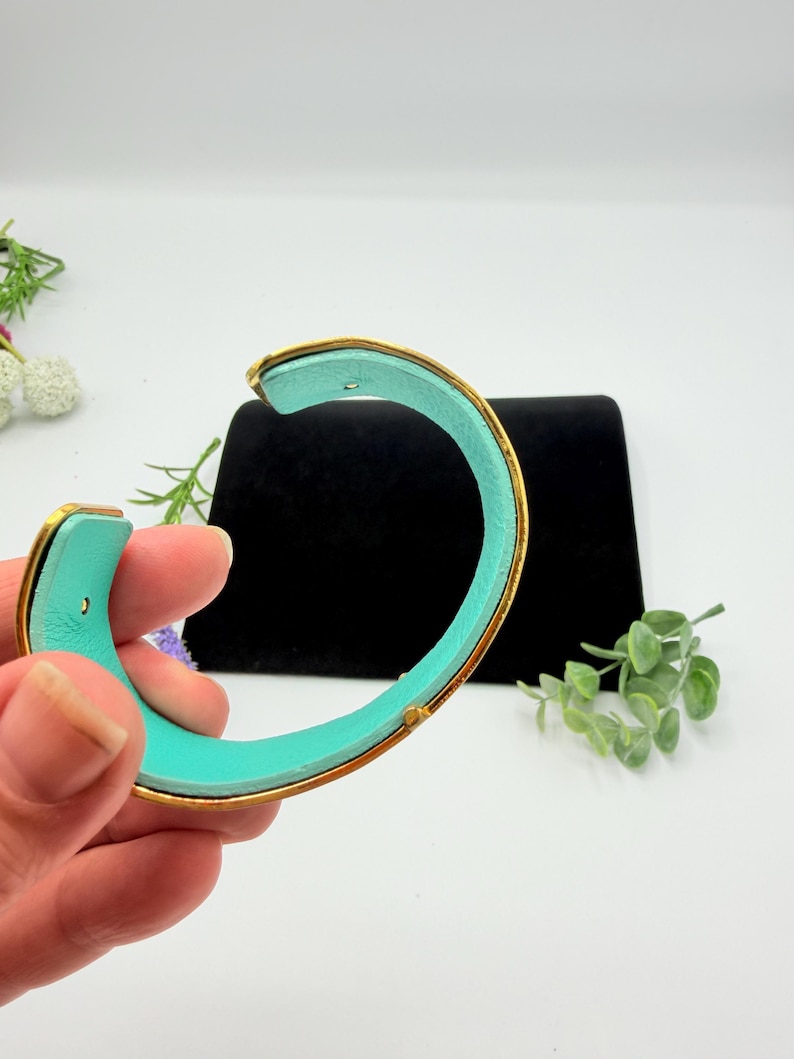 May include: A C-shaped cuff bracelet featuring a bright turquoise interior lining and a metallic gold-toned exterior frame. The bracelet has a smooth, curved design with a visible opening, showcasing a vibrant contrast between the inner colour and the outer gold-toned metal finish.
