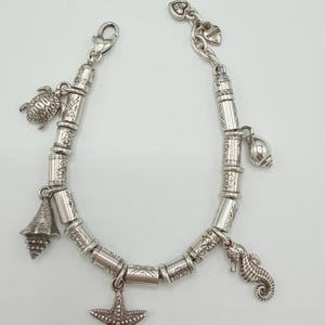 May include: A silver-toned charm bracelet featuring cylindrical beads with etched patterns and various sea-themed charms, including a sea turtle, a starfish, a seahorse, and two different types of seashells, secured with a lobster claw clasp.