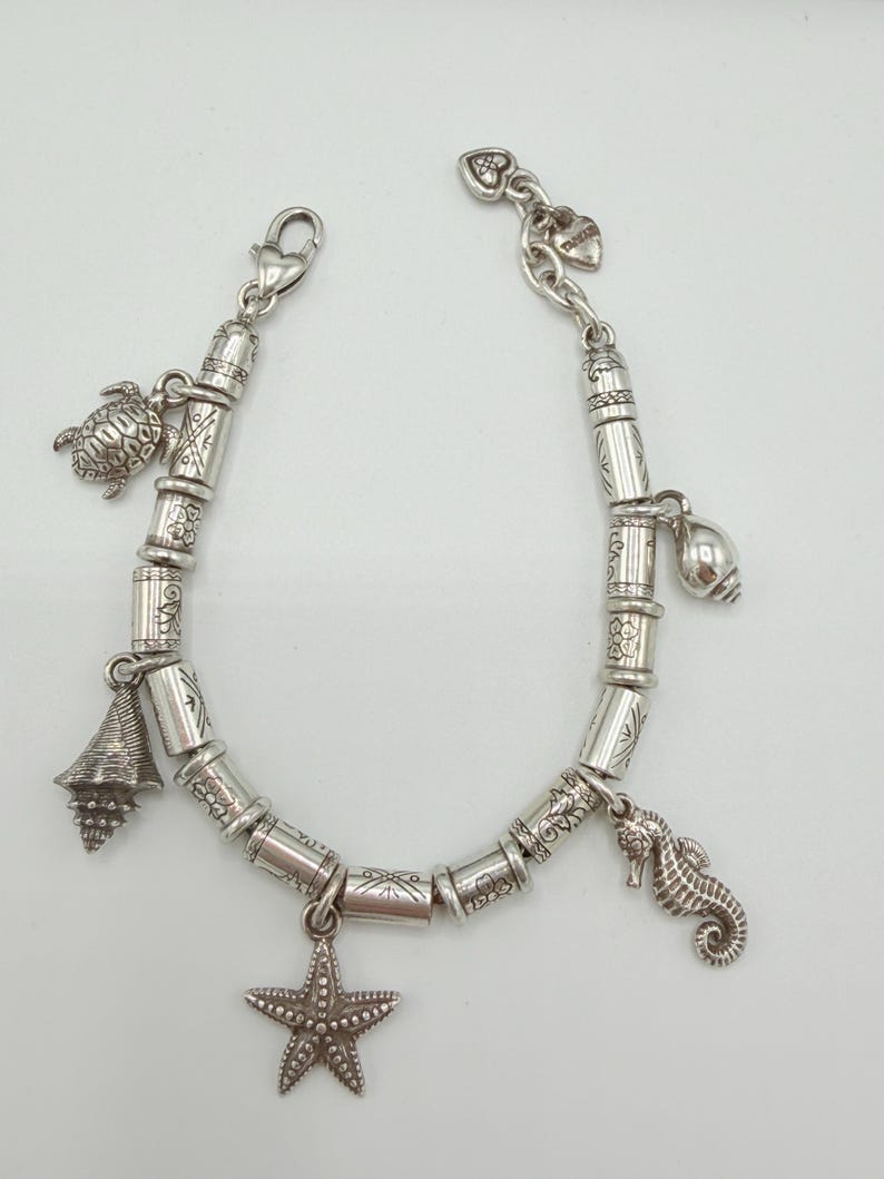 May include: A silver-toned charm bracelet featuring cylindrical beads with etched patterns and various sea-themed charms, including a sea turtle, a starfish, a seahorse, and two different types of seashells, secured with a lobster claw clasp.
