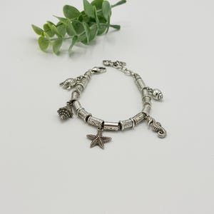 May include: A silver charm bracelet featuring a series of cylindrical metal beads with etched patterns. The bracelet includes dangling charms shaped like a starfish, a seahorse, a sea turtle, and two different types of seashells.
