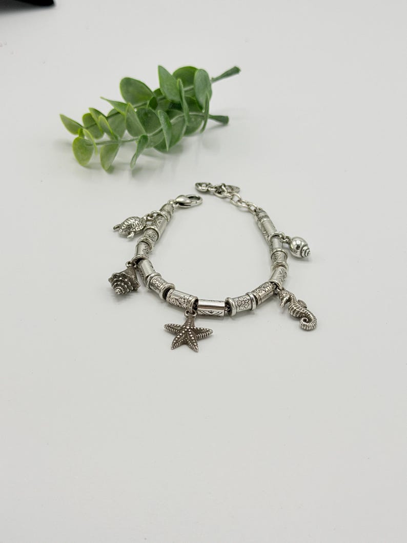May include: A silver charm bracelet featuring a series of cylindrical metal beads with etched patterns. The bracelet includes dangling charms shaped like a starfish, a seahorse, a sea turtle, and two different types of seashells.