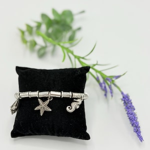 May include: A silver-toned charm bracelet featuring a series of cylindrical beads with etched patterns, interspersed with three dangling charms: a seashell, a five-pointed starfish, and a seahorse, all displayed on a black velvet cushion.