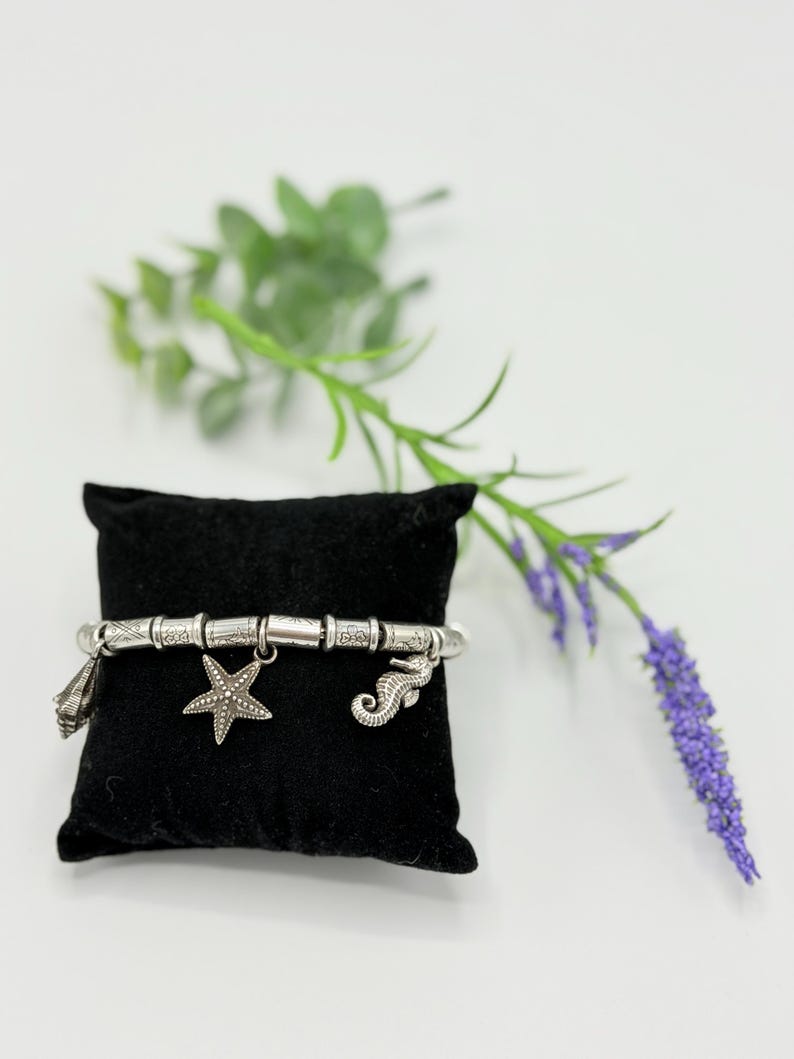 May include: A silver-toned charm bracelet featuring a series of cylindrical beads with etched patterns, interspersed with three dangling charms: a seashell, a five-pointed starfish, and a seahorse, all displayed on a black velvet cushion.