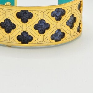 May include: A gold-tone cuff bracelet featuring a repeating lattice pattern with dark blue, textured, clover-shaped inlays. The interior of the bracelet is lined with a bright turquoise material, and the metal surface has a polished finish.