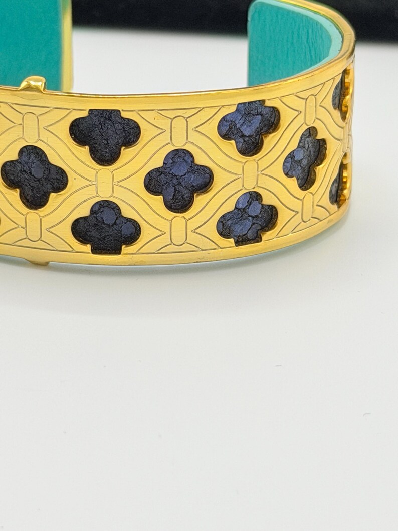 May include: A gold-tone cuff bracelet featuring a repeating lattice pattern with dark blue, textured, clover-shaped inlays. The interior of the bracelet is lined with a bright turquoise material, and the metal surface has a polished finish.