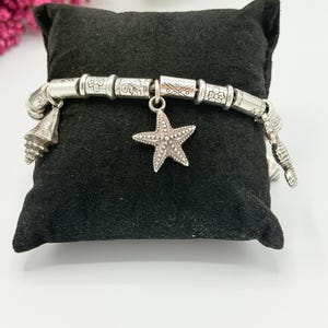 May include: A silver-toned charm bracelet featuring a series of cylindrical beads with engraved floral and geometric patterns. The bracelet includes dangling charms shaped like a starfish and various seashells, all arranged along the metal chain.