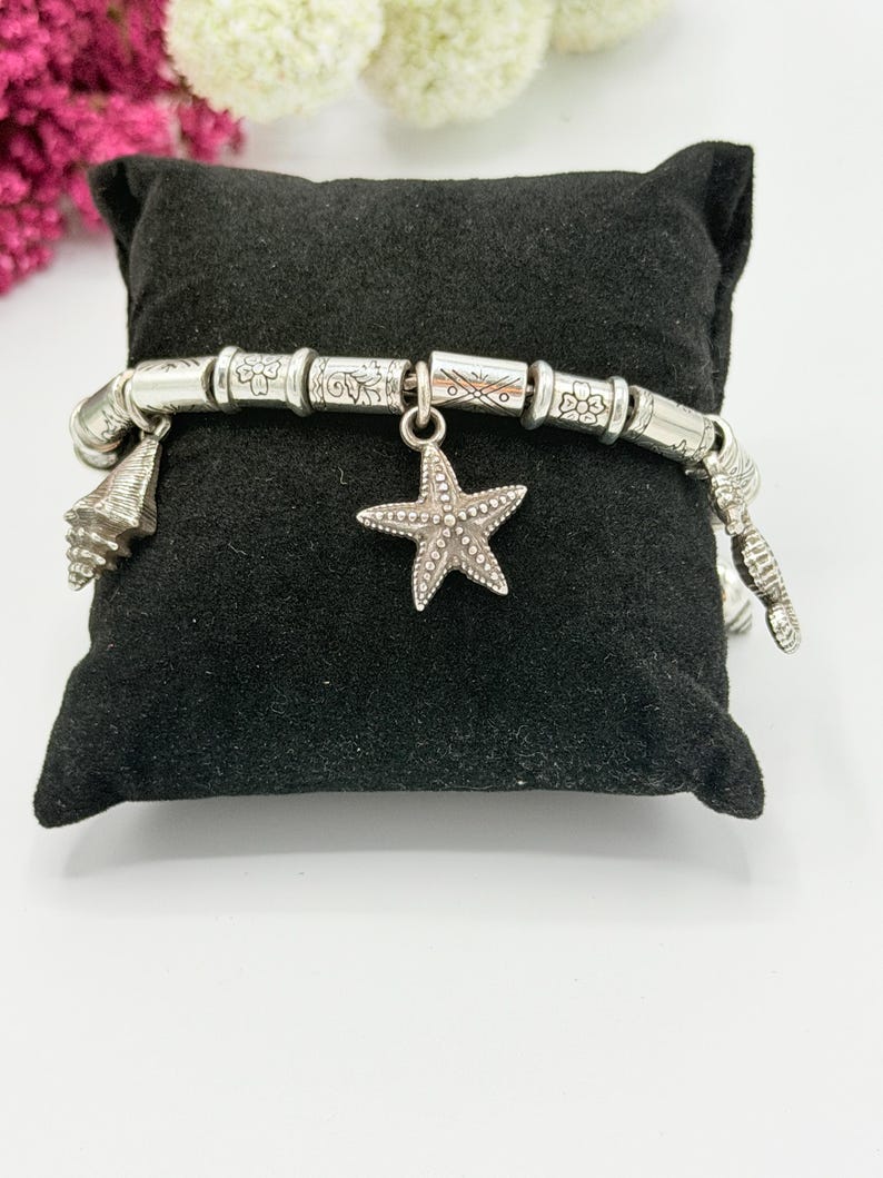 May include: A silver-toned charm bracelet featuring a series of cylindrical beads with engraved floral and geometric patterns. The bracelet includes dangling charms shaped like a starfish and various seashells, all arranged along the metal chain.