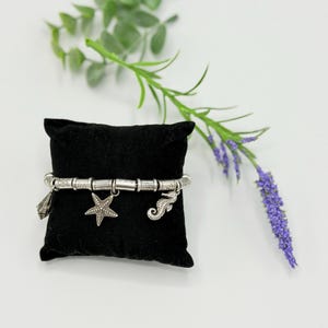 May include: A silver-toned charm bracelet featuring a series of textured cylindrical beads and three dangling charms: a seashell, a five-pointed starfish, and a seahorse. The bracelet is displayed on a small, square black velvet pillow.