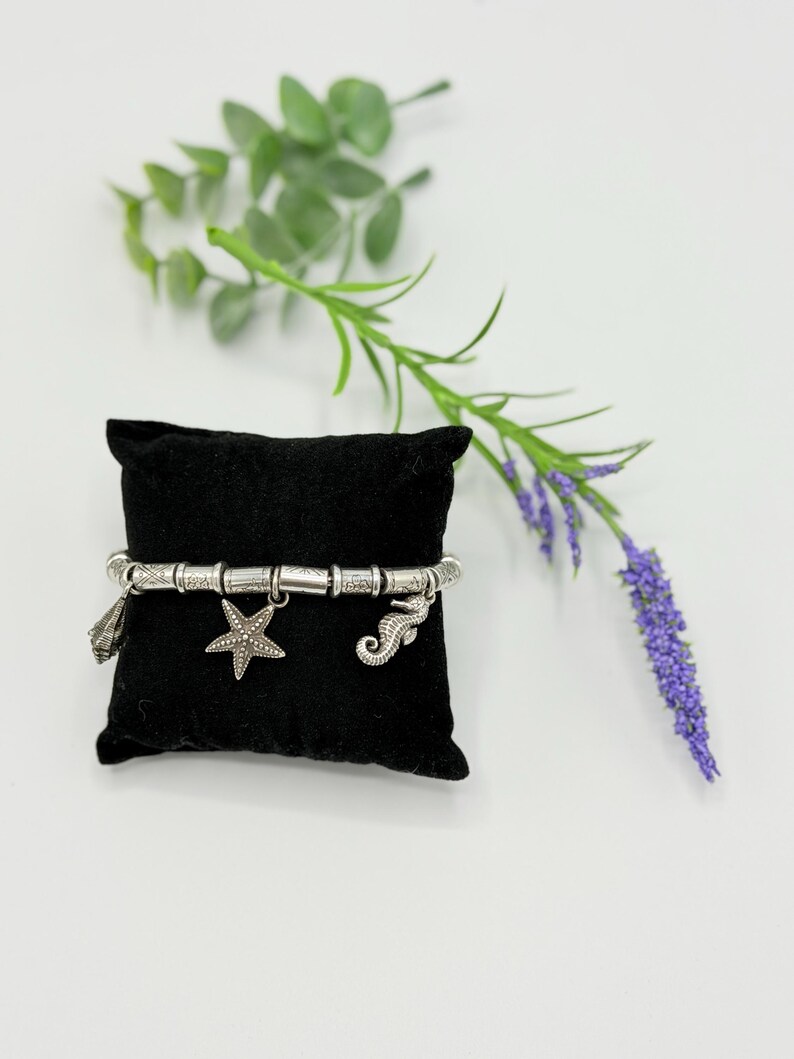 May include: A silver-toned charm bracelet featuring a series of textured cylindrical beads and three dangling charms: a seashell, a five-pointed starfish, and a seahorse. The bracelet is displayed on a small, square black velvet pillow.