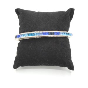 May include: A silver-toned bangle bracelet with a row of blue and teal square-cut gemstones. The bracelet is displayed on a black velvet pillow against a white background. The gemstones vary in shade from light teal to dark blue.