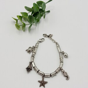 May include: A silver-toned charm bracelet featuring a series of cylindrical beads with etched patterns. The bracelet includes dangling charms shaped like a sea turtle, a conch shell, a starfish, and a seahorse, secured with a lobster claw clasp.