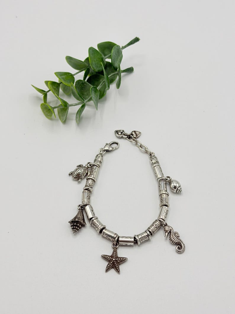 May include: A silver-toned charm bracelet featuring a series of cylindrical beads with etched patterns. The bracelet includes dangling charms shaped like a sea turtle, a conch shell, a starfish, and a seahorse, secured with a lobster claw clasp.