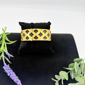 May include: A gold-toned cuff bracelet featuring a repeating quatrefoil cutout pattern. The metal framework overlays a dark blue textured material, creating a high-contrast design. The bracelet is displayed on a black velvet jewellery pillow.