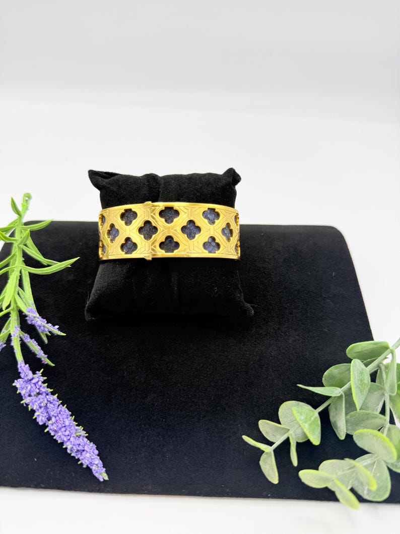 May include: A gold-toned cuff bracelet featuring a repeating quatrefoil cutout pattern. The metal framework overlays a dark blue textured material, creating a high-contrast design. The bracelet is displayed on a black velvet jewellery pillow.