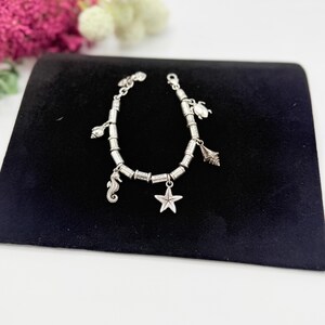 May include: A silver-toned charm bracelet featuring a series of cylindrical beads and multiple ocean-themed charms, including a seahorse, a starfish, a sea turtle, and various seashells, secured with a lobster claw clasp closure.