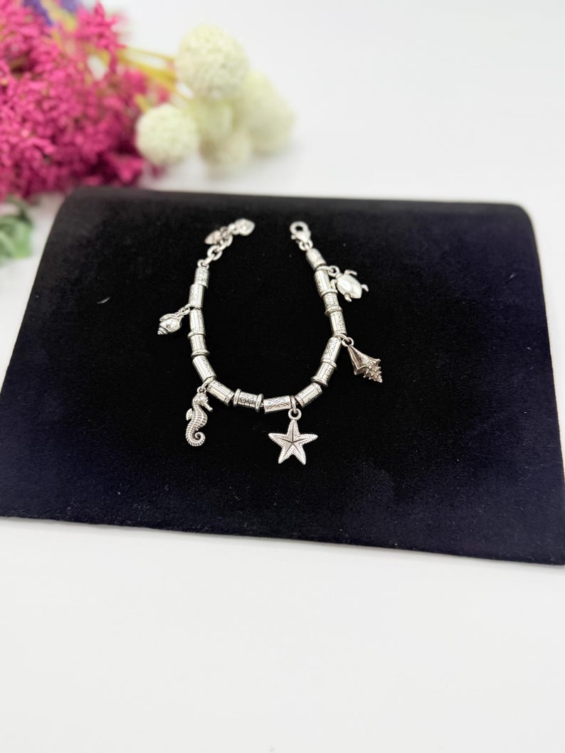 May include: A silver-toned charm bracelet featuring a series of cylindrical beads and multiple ocean-themed charms, including a seahorse, a starfish, a sea turtle, and various seashells, secured with a lobster claw clasp closure.