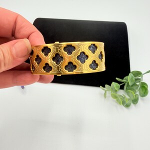 May include: A gold-toned cuff bracelet featuring a repeating quatrefoil cutout pattern. The openwork metal design reveals a dark, textured, blue-grey inlay beneath the surface, creating a high-contrast look across the entire band of the jewellery.