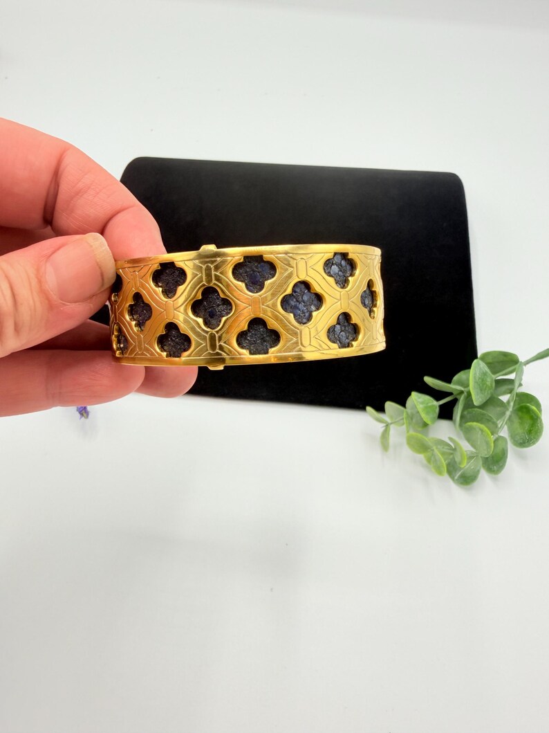 May include: A gold-toned cuff bracelet featuring a repeating quatrefoil cutout pattern. The openwork metal design reveals a dark, textured, blue-grey inlay beneath the surface, creating a high-contrast look across the entire band of the jewellery.