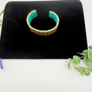May include: A gold-toned cuff bracelet featuring an intricate, openwork lattice pattern across the exterior surface. The interior of the bracelet is lined with a vibrant, solid turquoise-coloured material, providing a distinct contrast to the metallic gold exterior.
