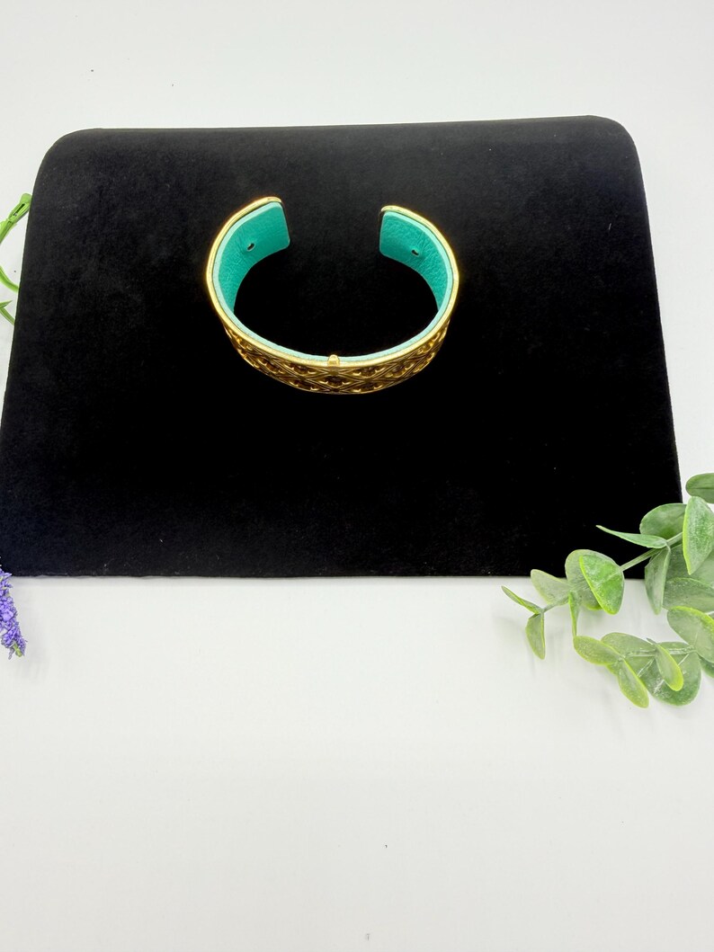 May include: A gold-toned cuff bracelet featuring an intricate, openwork lattice pattern across the exterior surface. The interior of the bracelet is lined with a vibrant, solid turquoise-coloured material, providing a distinct contrast to the metallic gold exterior.