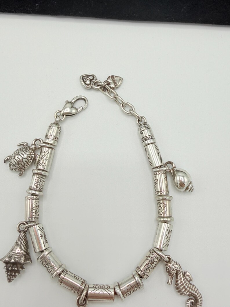 May include: A silver-toned charm bracelet featuring a series of cylindrical beads with engraved floral and geometric patterns. The bracelet includes dangling charms shaped like a sea turtle, a conch shell, and a seahorse, finished with a lobster clasp.