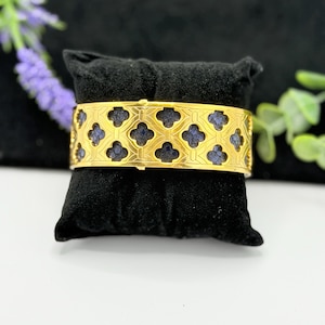 May include: A wide gold-toned cuff bracelet featuring a repeating cutout pattern of four-leaf clover shapes. The cutouts reveal a dark blue, textured inlay beneath the metallic surface, creating a high-contrast design across the entire band.