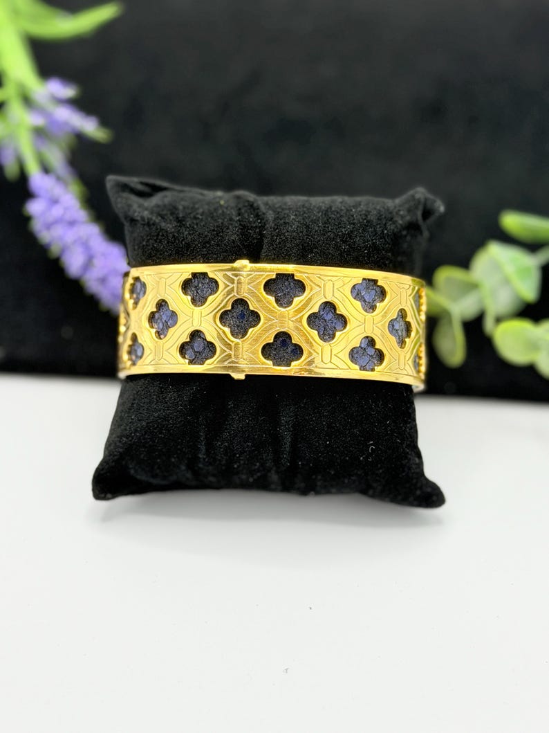 May include: A wide gold-toned cuff bracelet featuring a repeating cutout pattern of four-leaf clover shapes. The cutouts reveal a dark blue, textured inlay beneath the metallic surface, creating a high-contrast design across the entire band.
