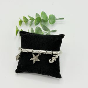 May include: A silver-toned charm bracelet featuring a series of cylindrical beads and three distinct dangling charms: a seashell, a five-pointed starfish, and a seahorse. The bracelet is displayed wrapped around a small, square black velvet pillow.