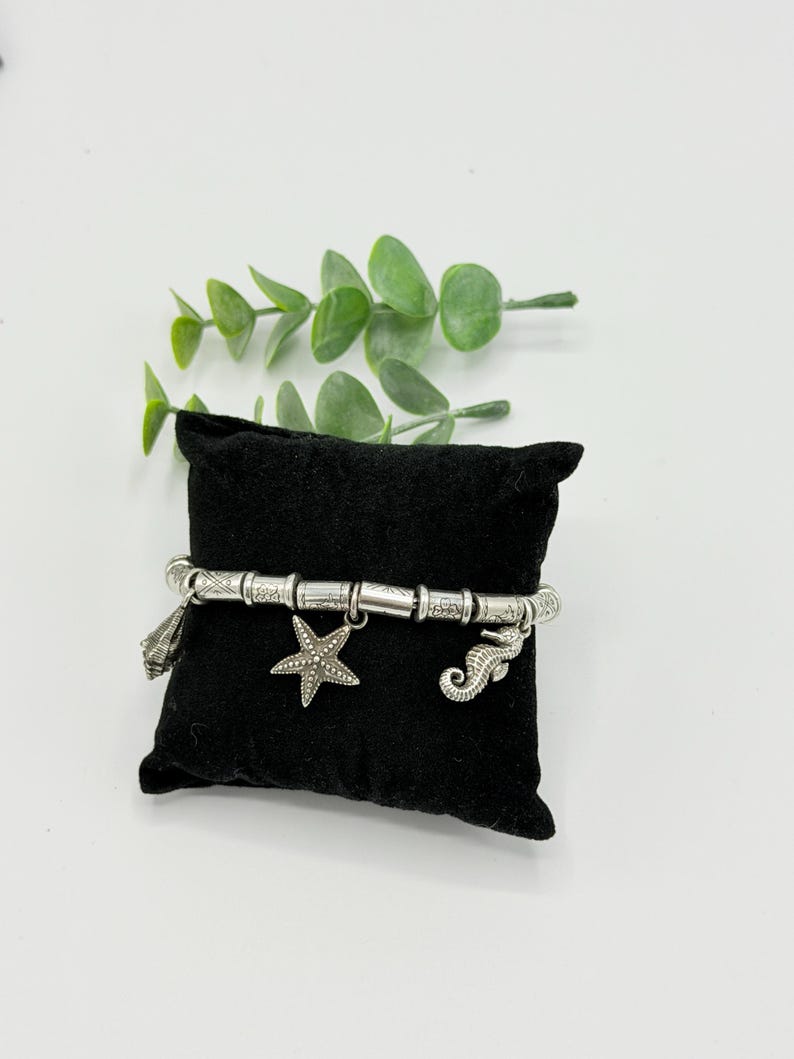 May include: A silver-toned charm bracelet featuring a series of cylindrical beads and three distinct dangling charms: a seashell, a five-pointed starfish, and a seahorse. The bracelet is displayed wrapped around a small, square black velvet pillow.