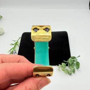 May include: A gold-tone metal cuff bracelet featuring a bright turquoise leather inlay. The top section displays a cutout clover pattern with dark stone accents and the word Brighton engraved in script. The design includes a hinged mechanism.